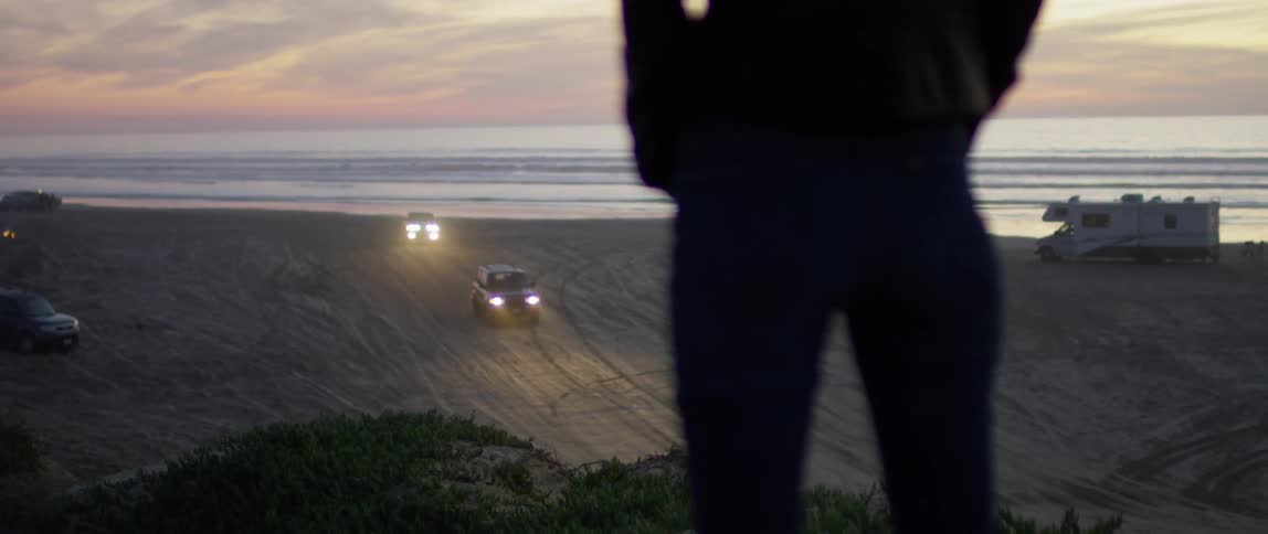 Person stands on a hill watching two vehicles drive on the sandy beach below
