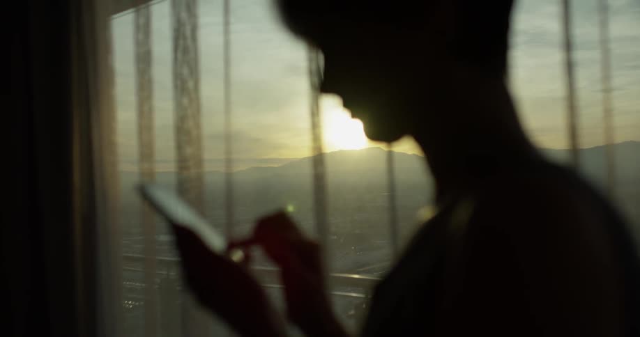 A woman uses her phone next to a window at sunset