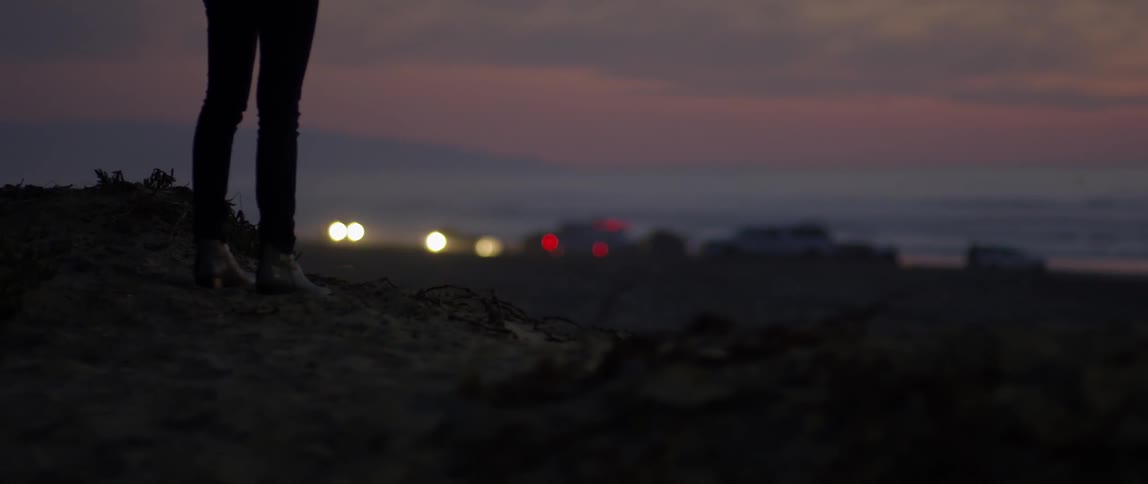 Woman walks in a field as a car drives nearby with its headlights on