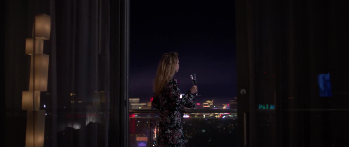 A woman takes a drink looking at the city from her balcony