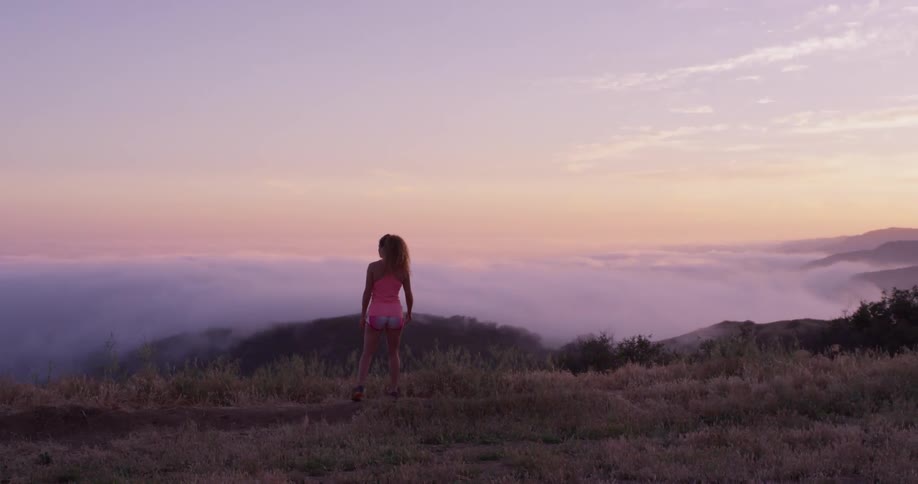 A woman walks along a grassy ridge then stops to look at the fog