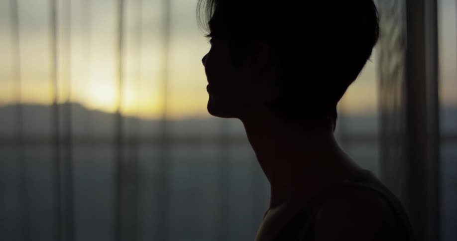 A woman stands next to a window at sunset
