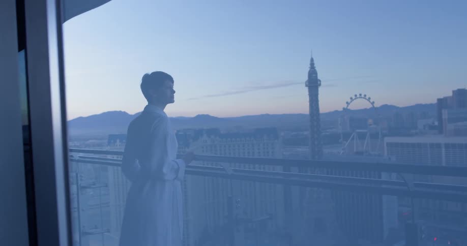 Woman in a white bathrobe stands on a balcony and looks out at the city