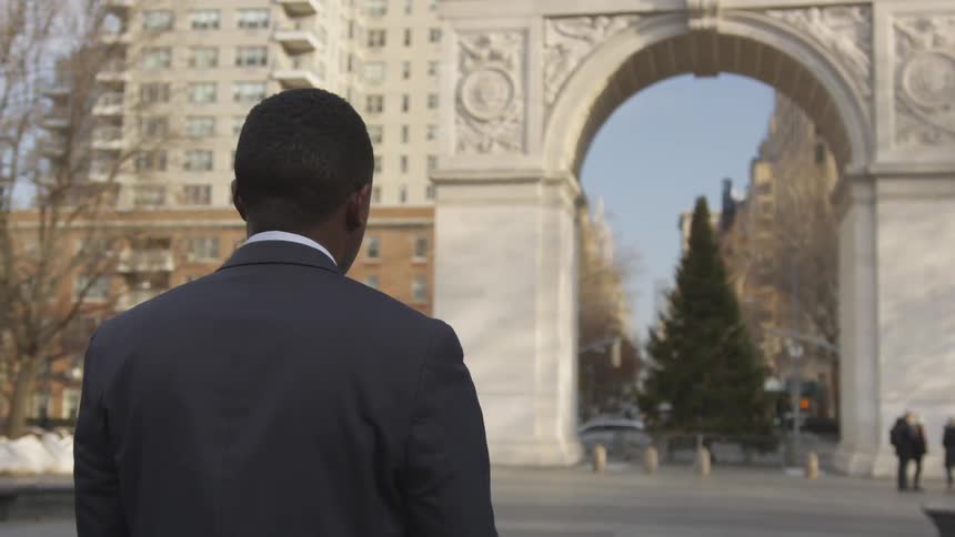 A man stands in a city plaza looking at an arch