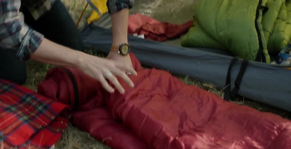 A brunette man wearing a red baseball cap talking and rolling up a sleeping bag