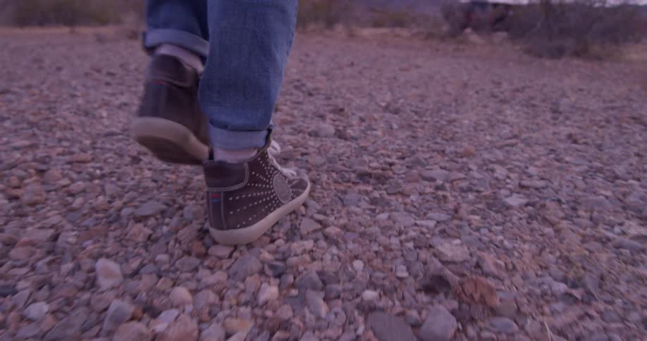 The legs and feet of a person walking over rocks and brush toward a car