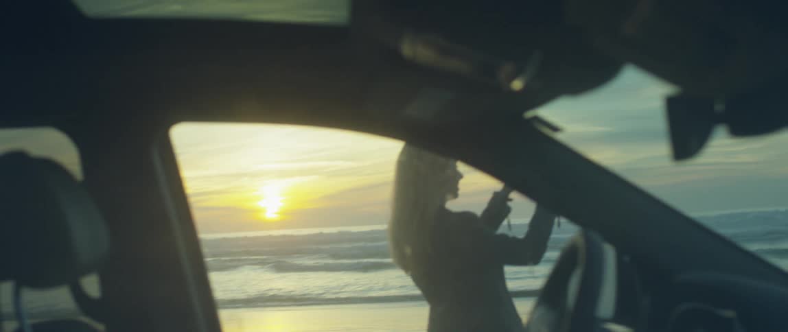 A woman stands outside her car and photographs the ocean sunset