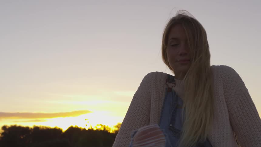 A woman sits outside at sunset with trees behind her
