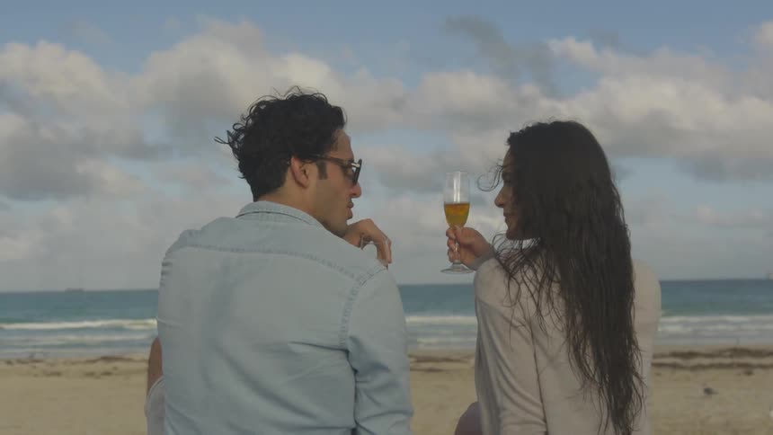 A couple are clinking their champagne glasses together as they sit on a beach