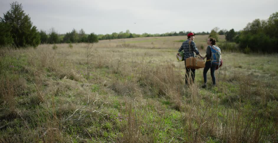 A man and woman walking through a field while carrying duffel bags