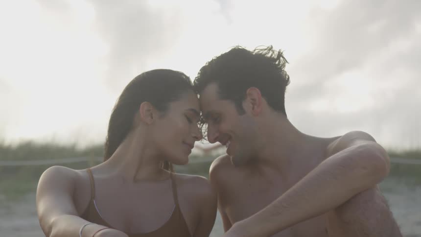 A man and woman sitting on the beach smiling and resting their heads together on a cloudy day