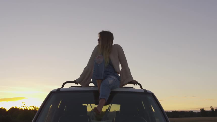 A woman is sitting on the car at sunset