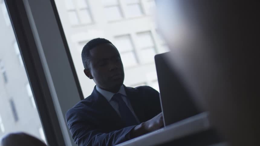A man sits at a desk working on a laptop in front of a window with a large building outside