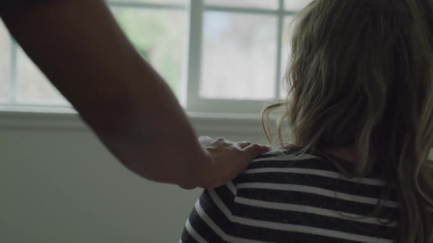 A woman sits in front of a window and pulls away when a man approaches and puts his hand on her shoulder