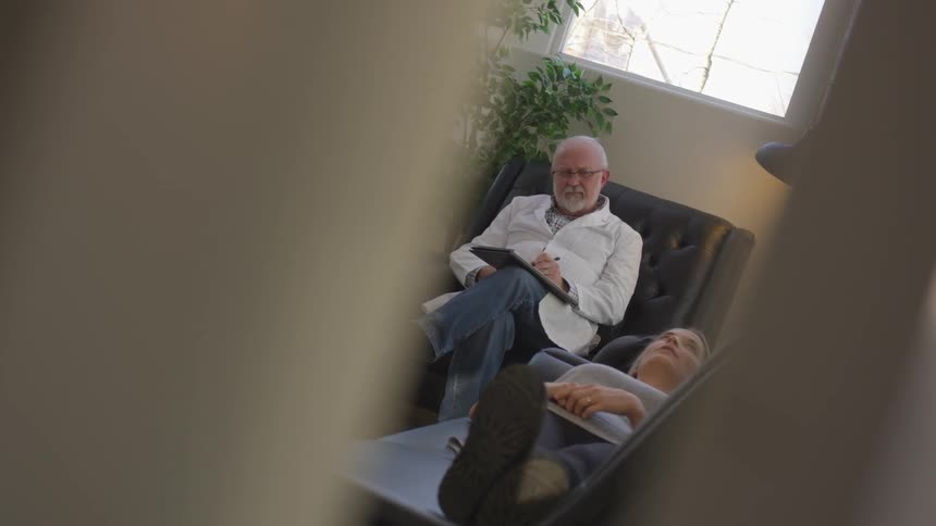 A man in a white lab coat sits on a chair in an office holding a pad and pen while a woman lays on a couch and talks