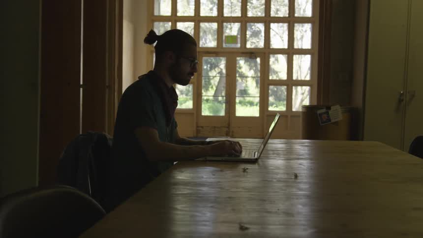 A man is sitting inside in daylight at a table using a laptop
