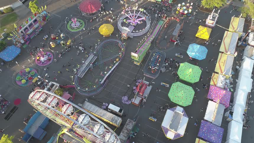 Many people walk through a large carnival with tents a ferris wheel a giant slide and other rides