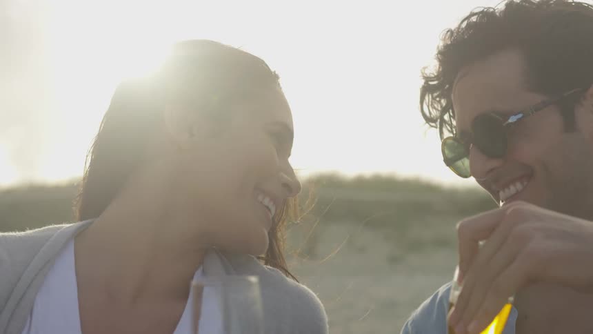 A man and a woman smile while holding champagne glasses on the beach on a partly cloudy day
