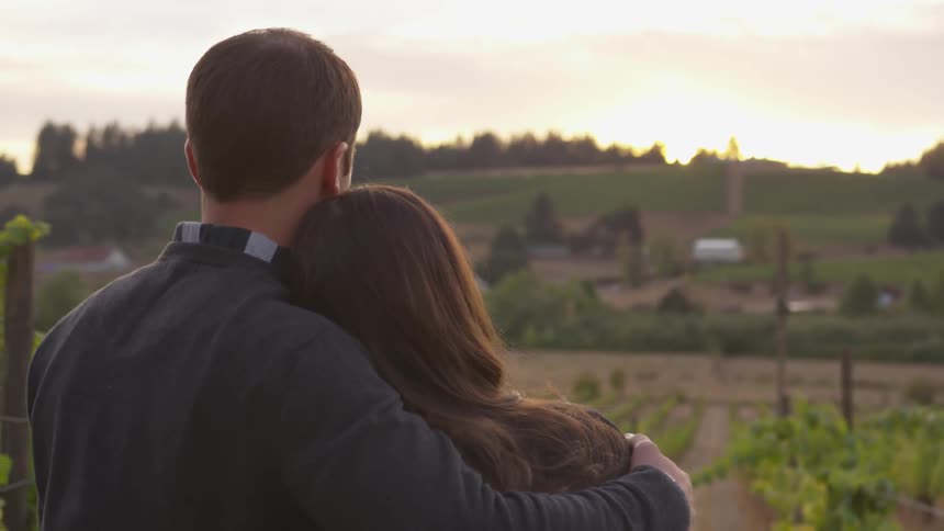A couple are leaning on each other as they take in a vineyard view at sunset