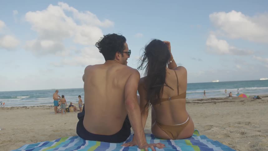 A couple in bathing suits sit on a towel at the beach