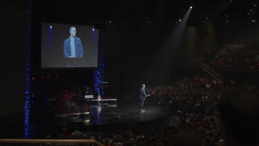 A man performs on stage as a teleprompter works behind him