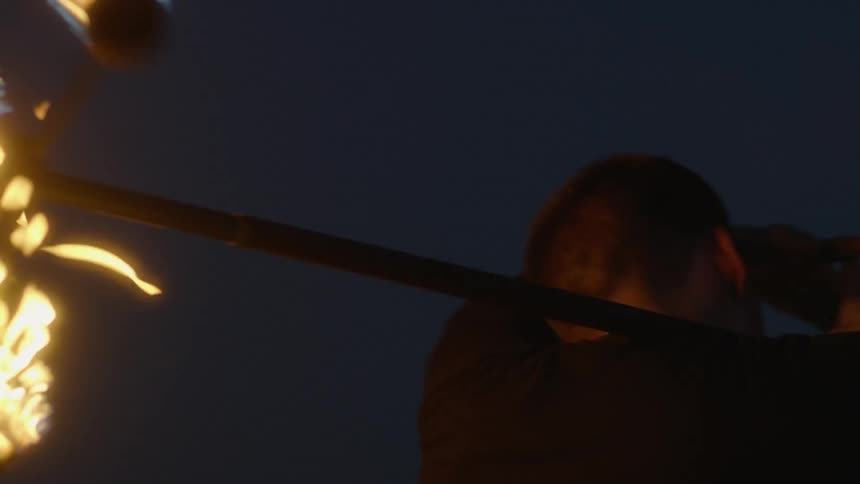 A performer is spinning torches on a bar outside at dusk
