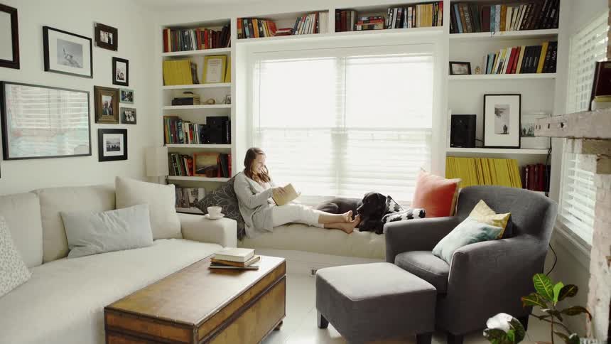 Woman sits on a couch next to a window reading a book