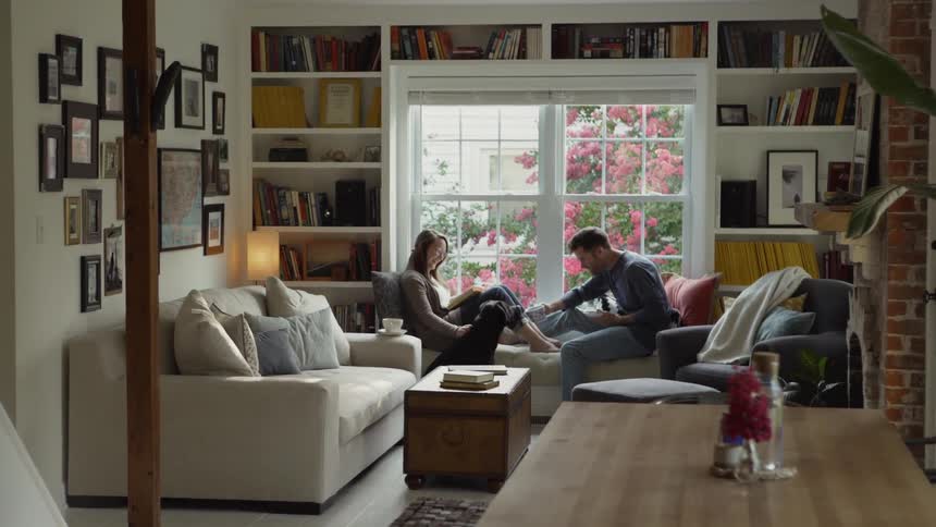 A man and woman sit in a window seat at day time while the man drinks from a teacup and the woman reads while petting a dog