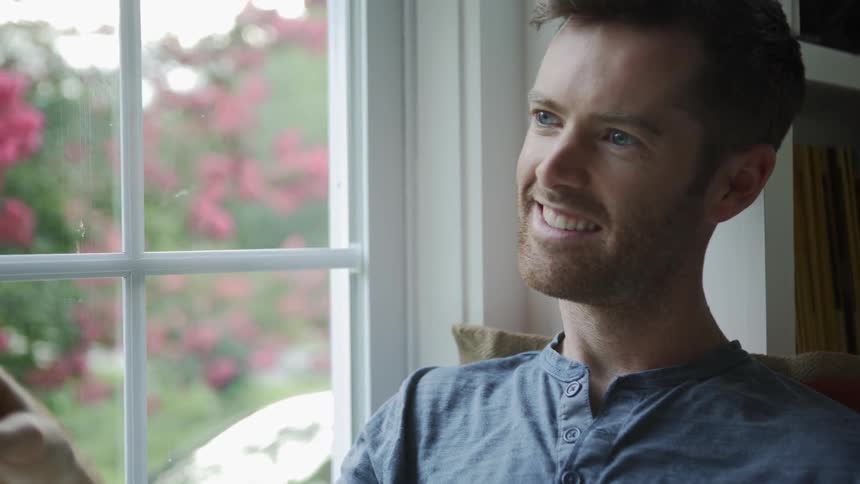 A man sits in a home in a window seat holding a book and smiling
