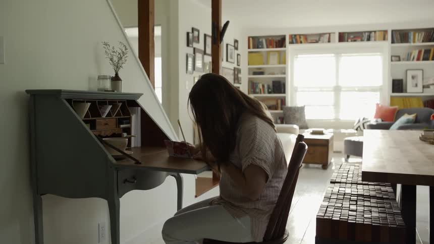 A woman sits at a secretary desk and writes in a dining room at day time