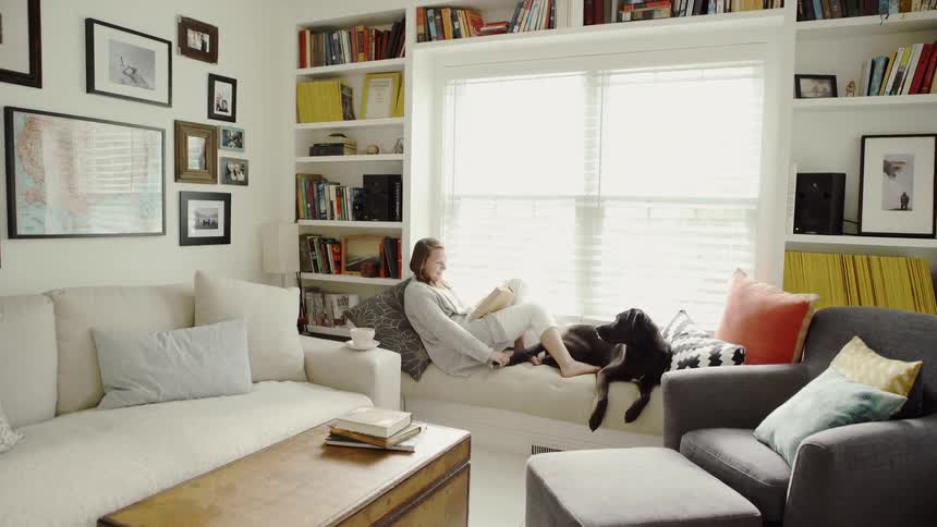Woman sits on a couch next to a window reading with a dog at her feet