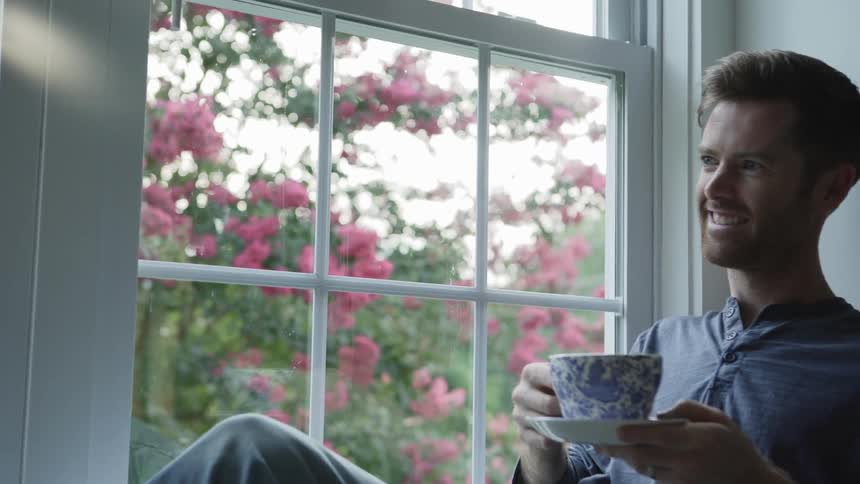 Man smiles next to a window holding a teacup