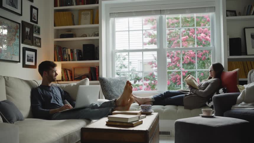 A man sits on a couch with his laptop while a woman reads at a window seat