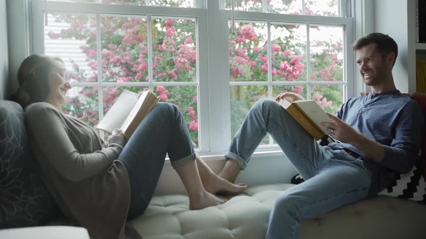 Man and woman touch their feet together as they sit on the sofa reading