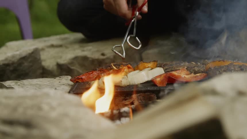 A person uses tongs to flip food on a flaming grill