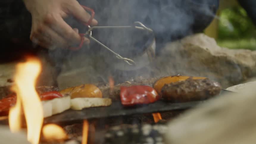 A man uses tongs to turn burgers on a grill with veggies outside