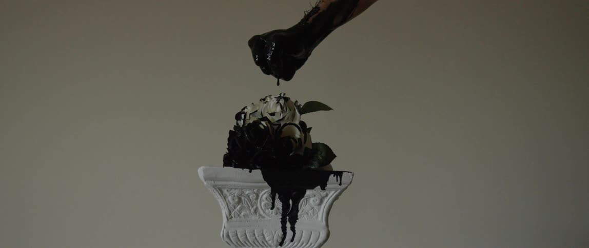 A hand covered in black ink drips down onto a white rose on a pedestal