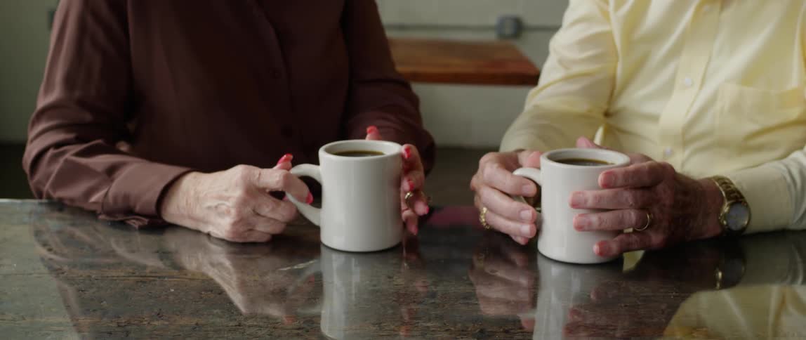 Two elderly women sit in a coffee shop with their hands shown holding cups