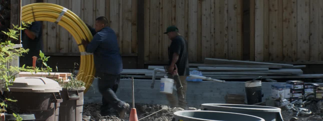 Two men at a construction site push a large spool of wires as a third man carrying a bucket and a pipe walks behind them