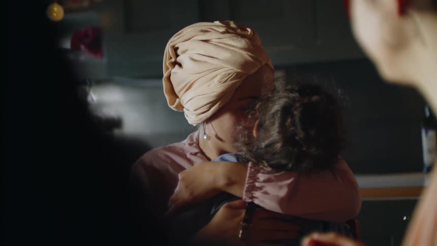A woman sitting in a dark dining area smiles as she hugs and kisses a little girl