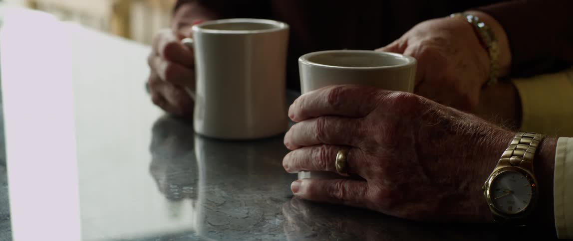 The hands of an elderly woman and elderly man shown holding coffee cups as she clasps his hand