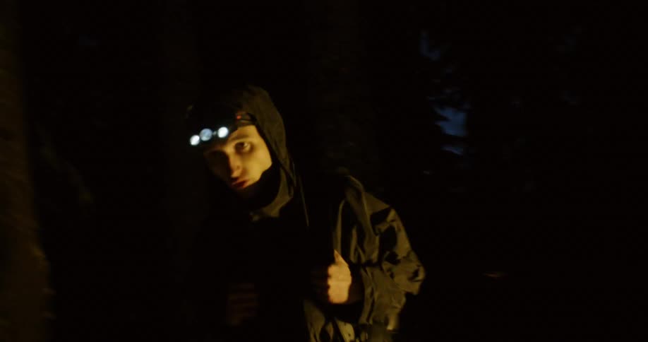 A man carrying a backpack and wearing a headlamp walks through a snowy forest at night