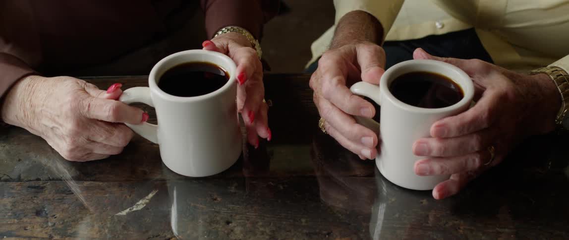 The hands of an elderly couple, each holding mugs of coffee