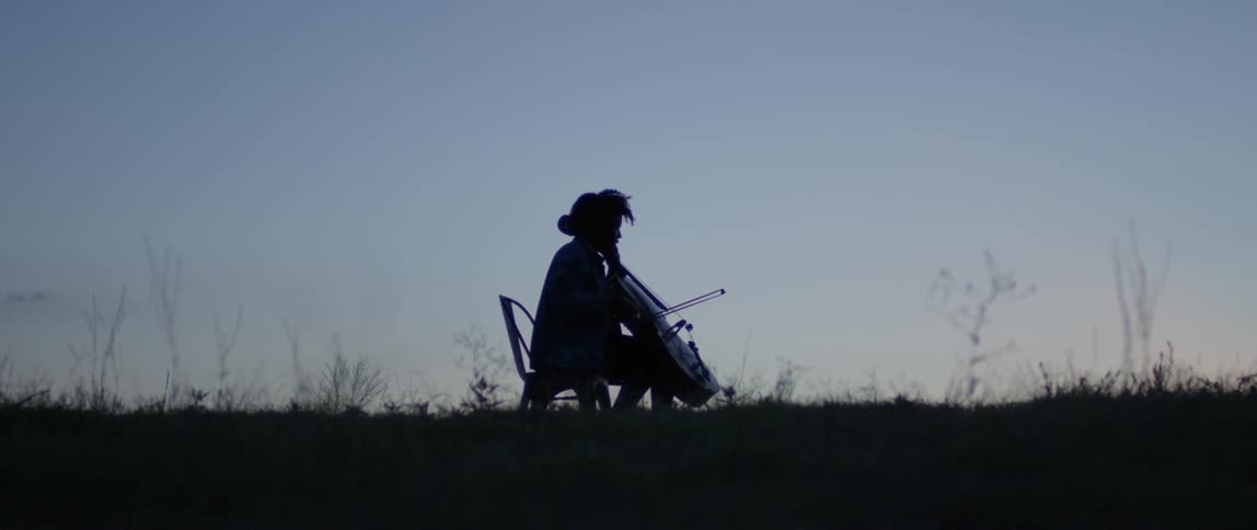 A person plays a cello sitting outside at sunset
