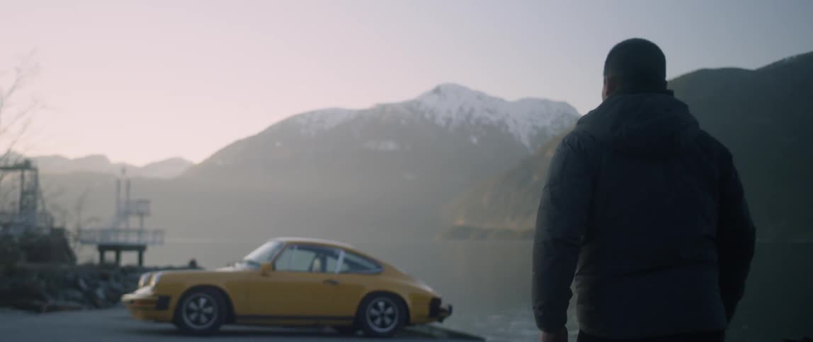 A man walks towards a yellow car parked next to a body of water at sunset with mountains in the background