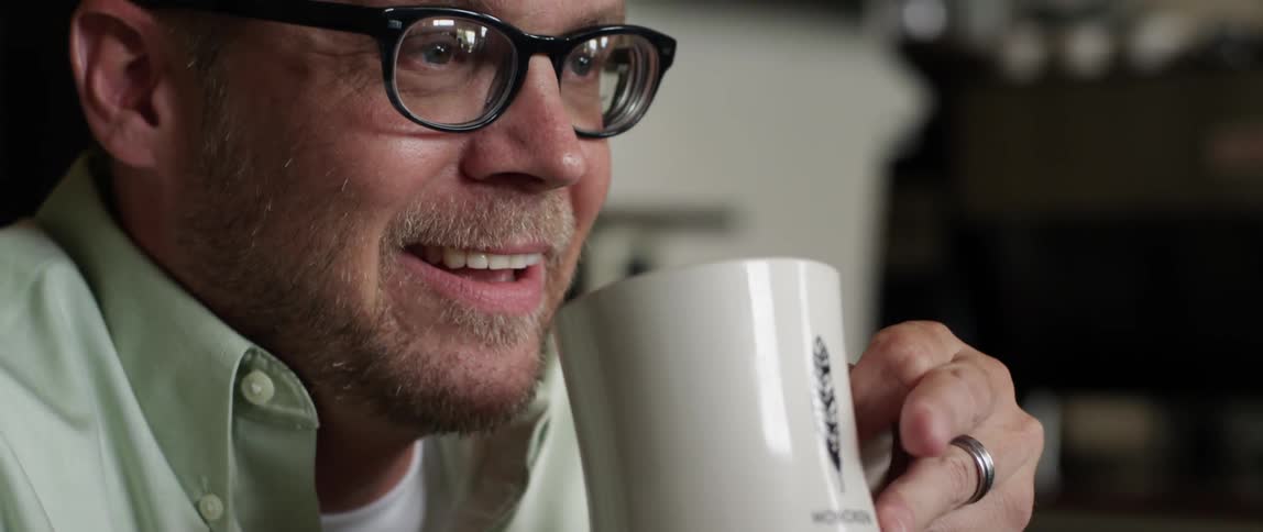 A man wearing glasses drinks from a white mug