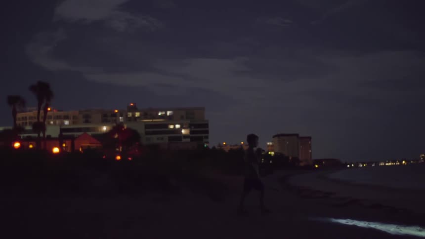 A boy walking on the beach with a flashlight in front of some buildings at night