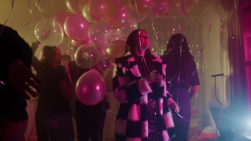 A group of men and woman dance and jump at a house party with flashing lights and balloons