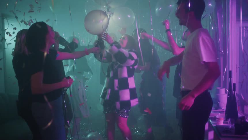 A crowd of men and women smile and dance in a dark room with flashing lights confetti and balloons