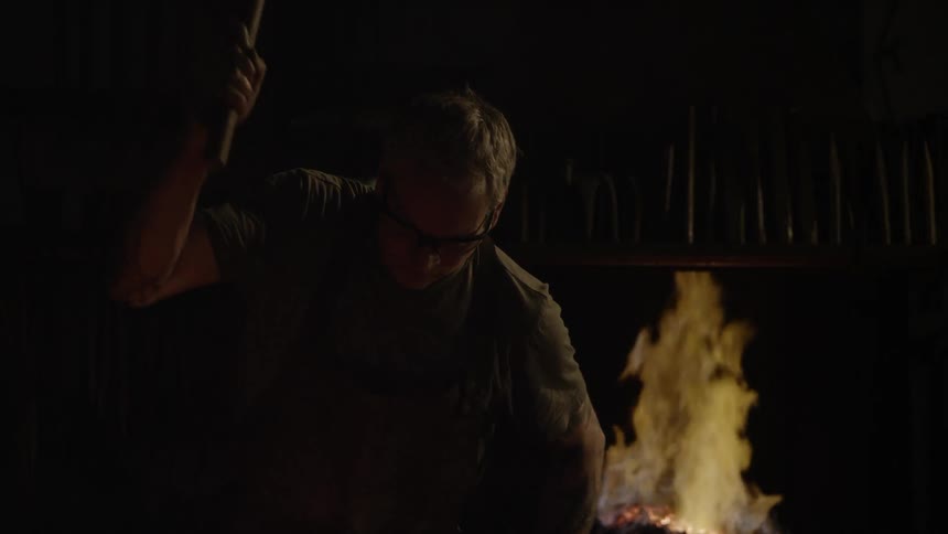 A man swings a mallet in the dark in front of a fire